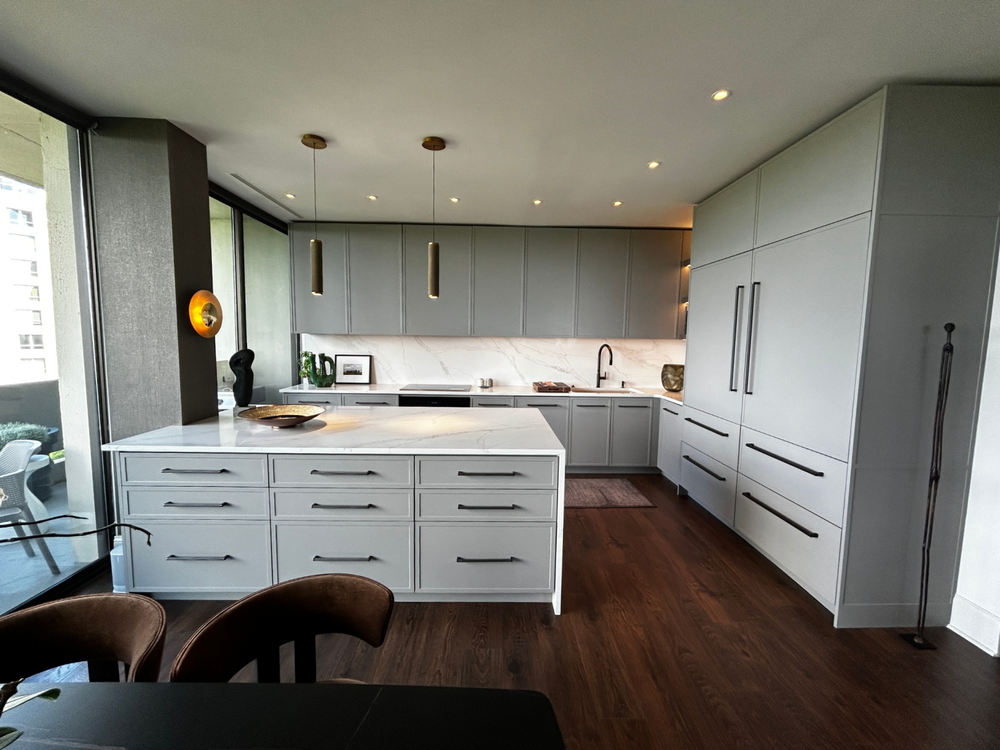 A modern minimalist kitchen with floor-to-ceiling light grey cabinets featuring black pulls, a white marble countertop, and a sleek island. The kitchen is illuminated by recessed lighting and pendant lights, with a marble backsplash extending along the wall. Large floor-to-ceiling windows offer a view of the outdoors, complemented by dark wooden flooring and stylish brown upholstered chairs around a black dining table. Decorative items include a golden wall sconce and a ceramic vase.
